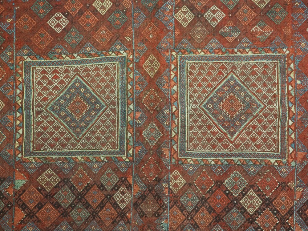 An antique Caucasian flatweave rug displaying geometric diamond medallions in rust red, navy blue, and cream wool with fringed ends. View IX