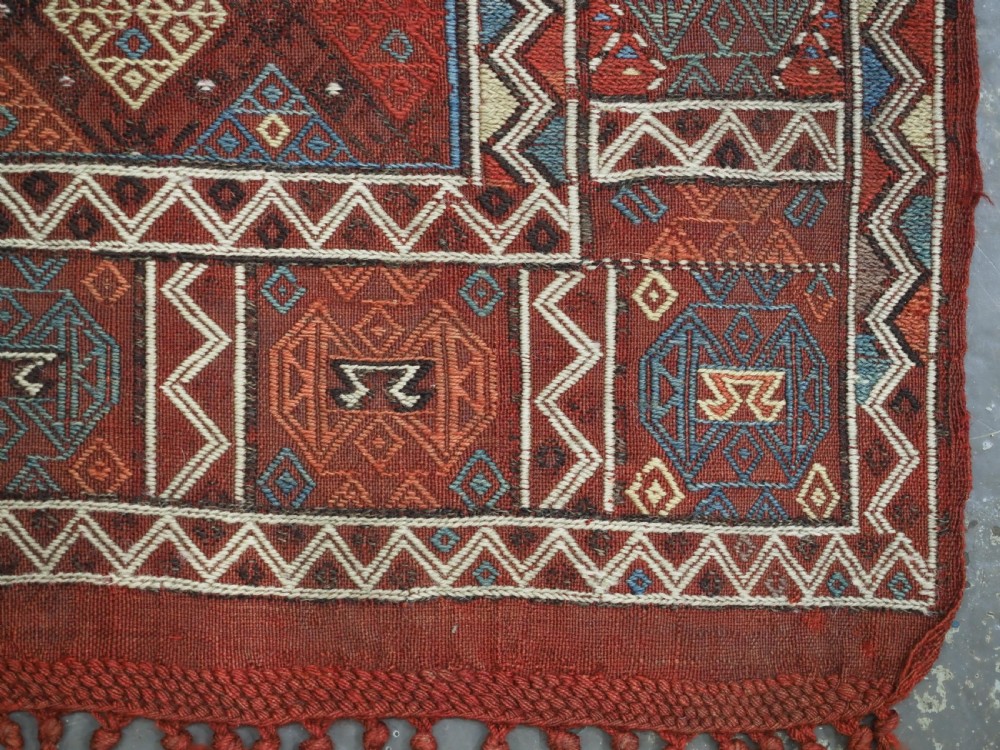 An antique Caucasian flatweave rug displaying geometric diamond medallions in rust red, navy blue, and cream wool with fringed ends. View X