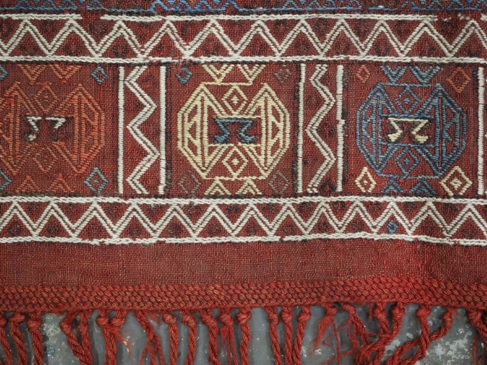 An antique Caucasian flatweave rug displaying geometric diamond medallions in rust red, navy blue, and cream wool with fringed ends. View XI