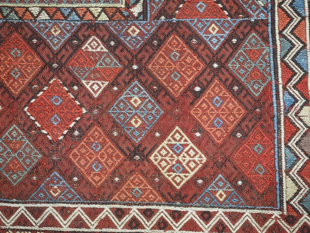 An antique Caucasian flatweave rug displaying geometric diamond medallions in rust red, navy blue, and cream wool with fringed ends. View XII
