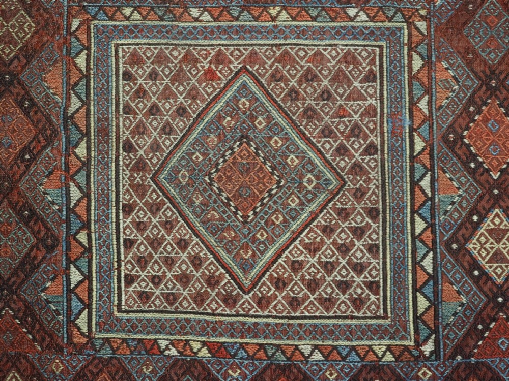 An antique Caucasian flatweave rug displaying geometric diamond medallions in rust red, navy blue, and cream wool with fringed ends. View XIII
