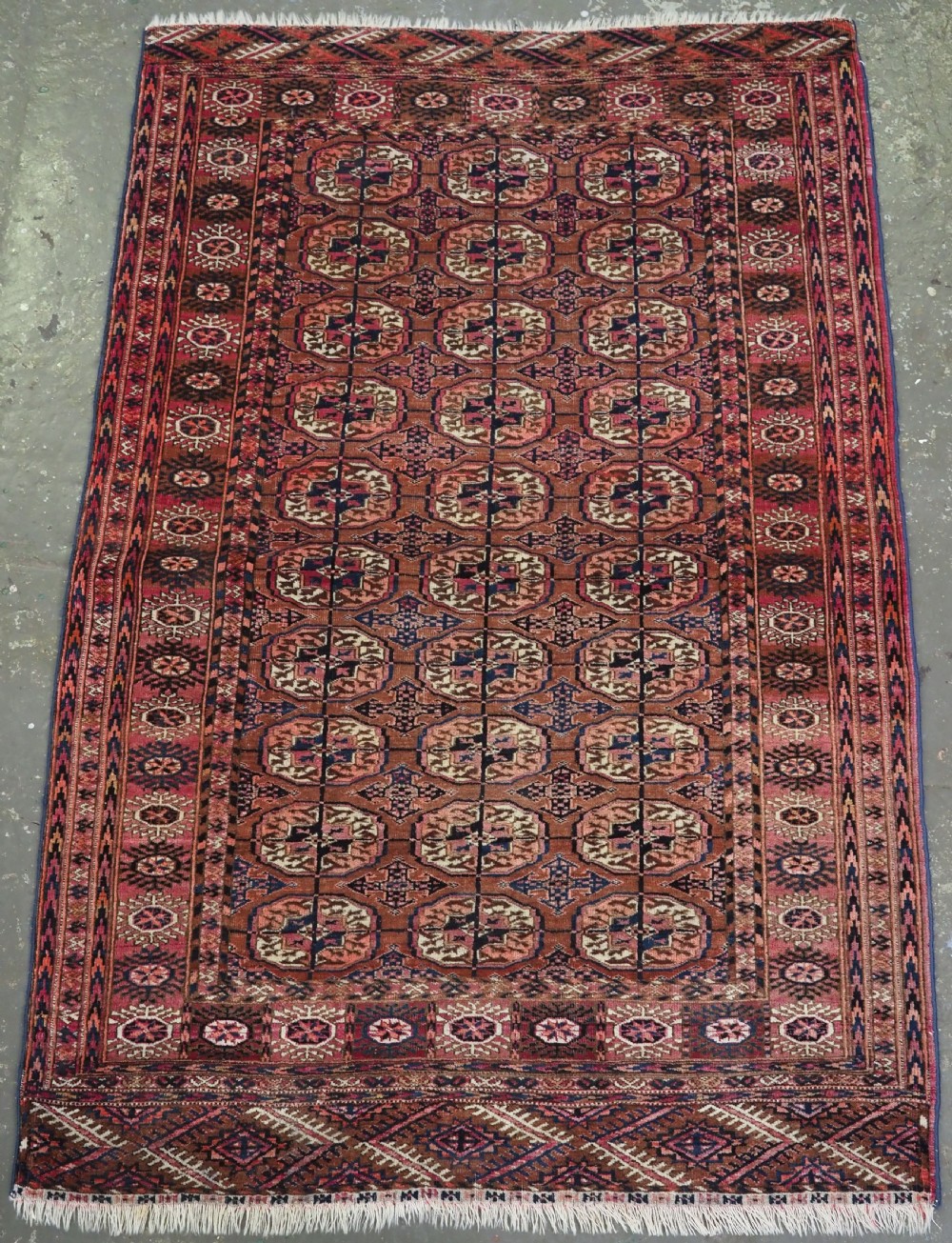 An antique rectangular hand-knotted wool Tekke Turkmen rug featuring repeating octagonal gul motifs in terracotta, navy, and cream tones with geometric borders.