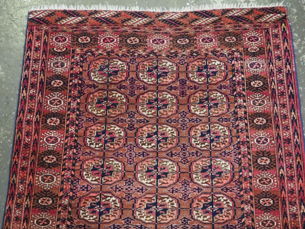 An antique rectangular hand-knotted wool Tekke Turkmen rug featuring repeating octagonal gul motifs in terracotta, navy, and cream tones with geometric borders. View III