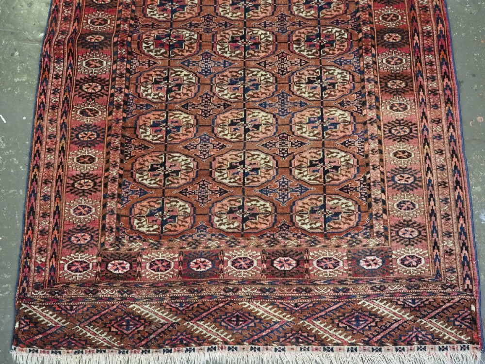 An antique rectangular hand-knotted wool Tekke Turkmen rug featuring repeating octagonal gul motifs in terracotta, navy, and cream tones with geometric borders. View IV
