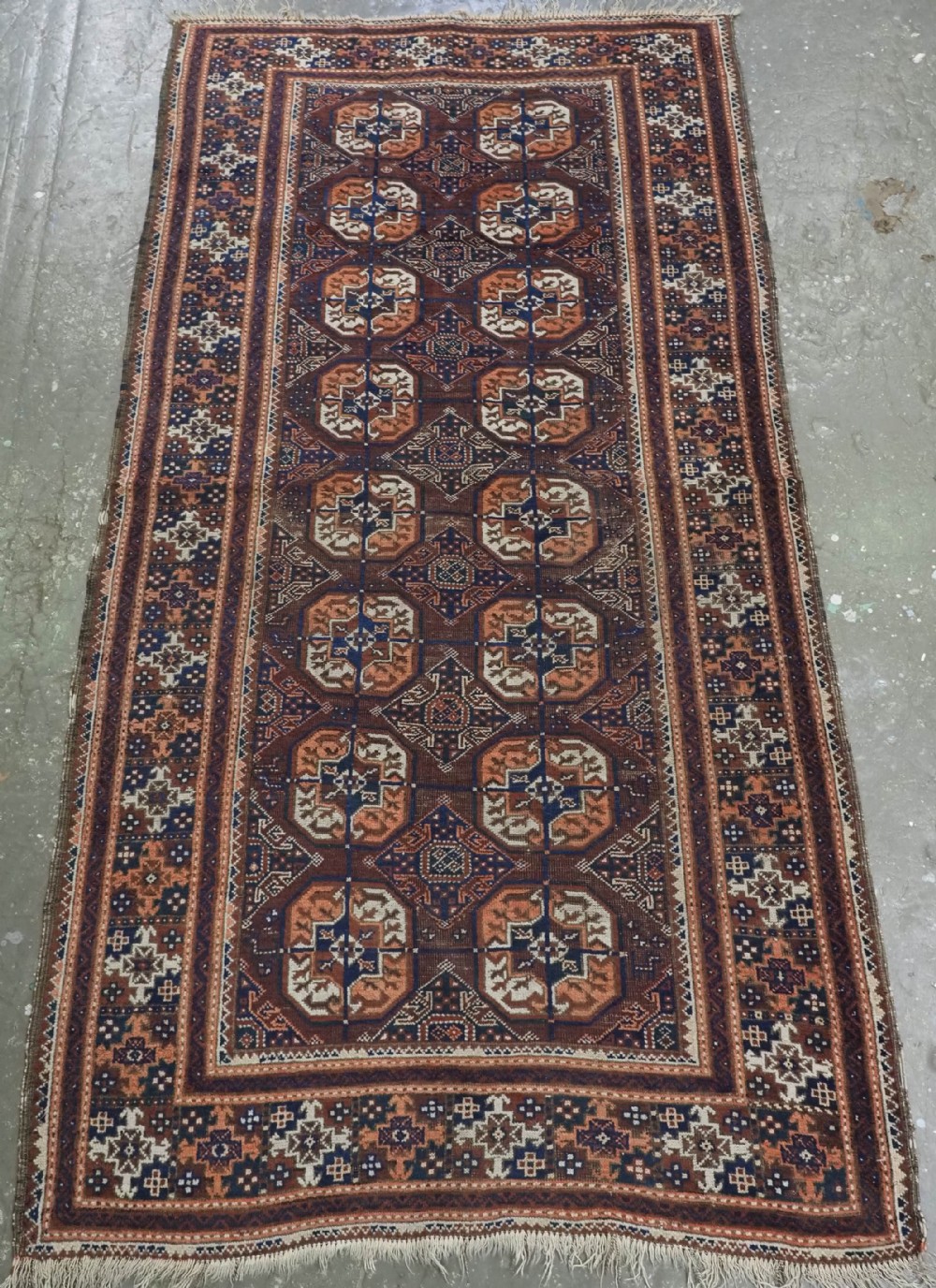 An antique hand-knotted Baluch wool rug featuring repeating geometric octagonal medallions in terracotta, navy, and ivory across a dark brown field with intricate multi-banded borders.