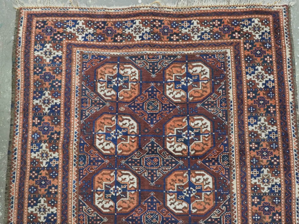 An antique hand-knotted Baluch wool rug featuring repeating geometric octagonal medallions in terracotta, navy, and ivory across a dark brown field with intricate multi-banded borders. View III