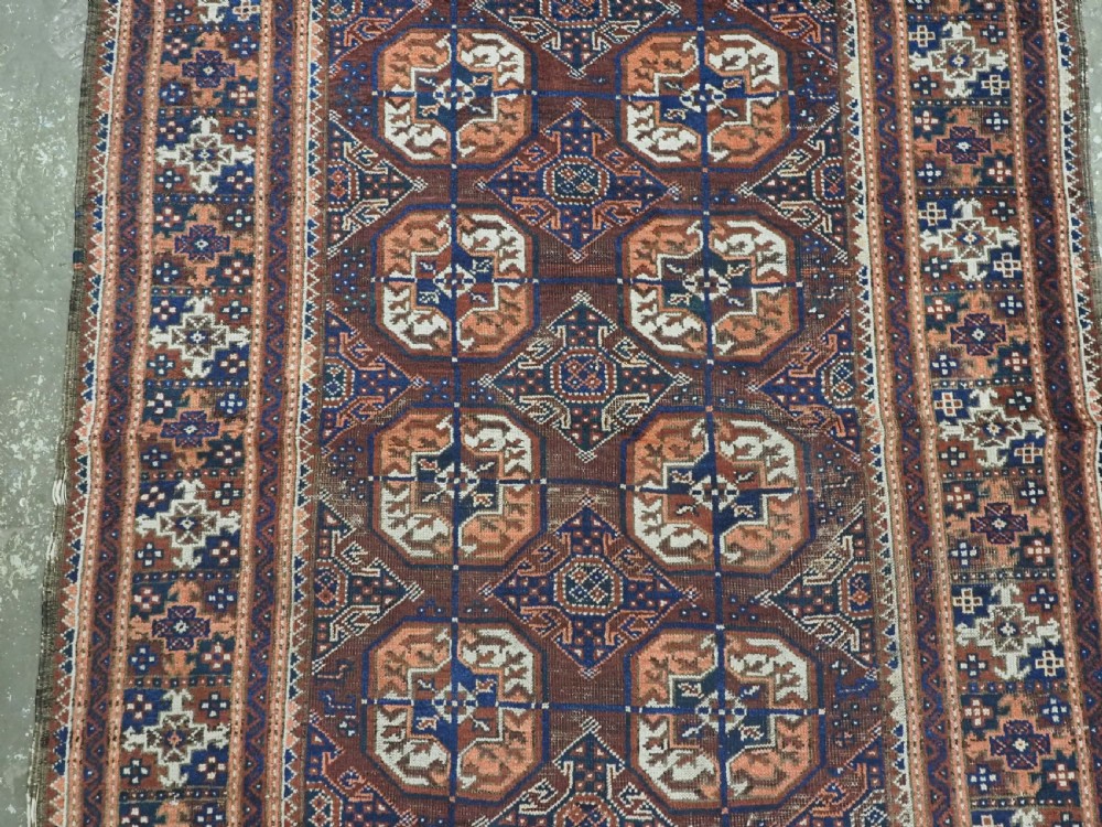 An antique hand-knotted Baluch wool rug featuring repeating geometric octagonal medallions in terracotta, navy, and ivory across a dark brown field with intricate multi-banded borders. View IV