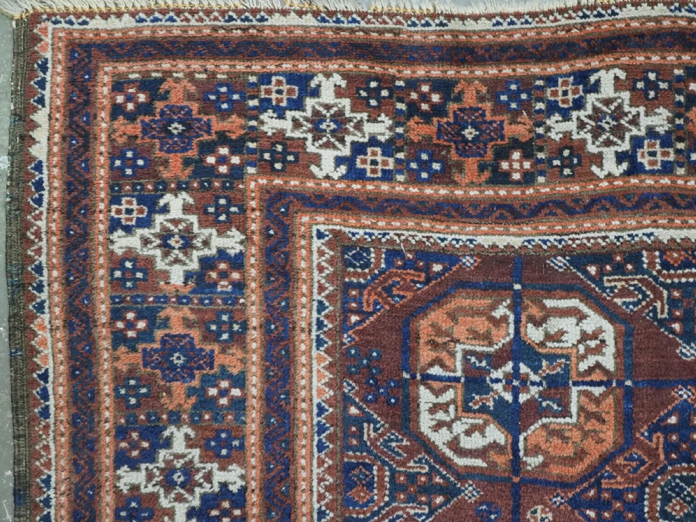 An antique hand-knotted Baluch wool rug featuring repeating geometric octagonal medallions in terracotta, navy, and ivory across a dark brown field with intricate multi-banded borders. View V