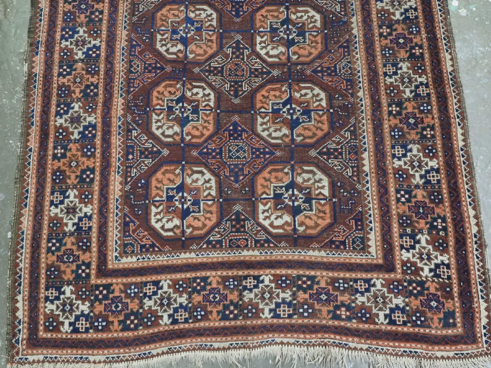 An antique hand-knotted Baluch wool rug featuring repeating geometric octagonal medallions in terracotta, navy, and ivory across a dark brown field with intricate multi-banded borders. View VI