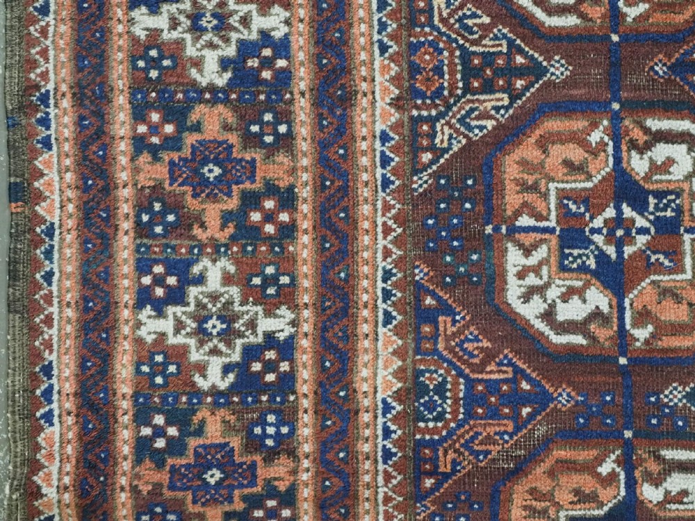 An antique hand-knotted Baluch wool rug featuring repeating geometric octagonal medallions in terracotta, navy, and ivory across a dark brown field with intricate multi-banded borders. View VII