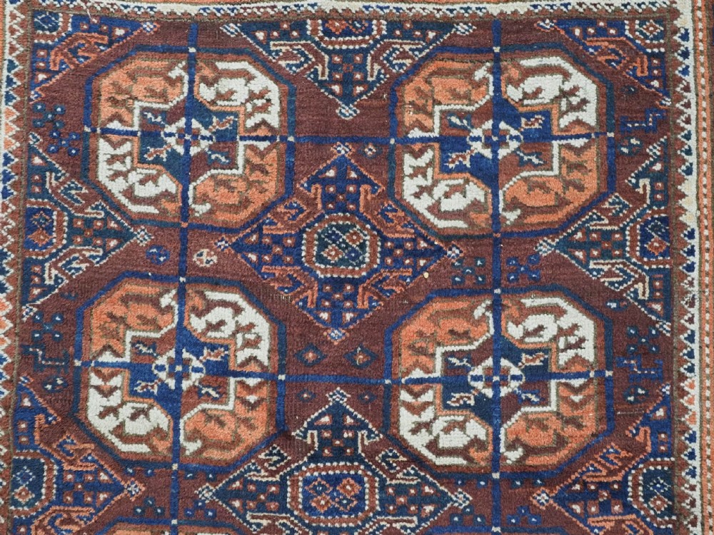 An antique hand-knotted Baluch wool rug featuring repeating geometric octagonal medallions in terracotta, navy, and ivory across a dark brown field with intricate multi-banded borders. View VIII