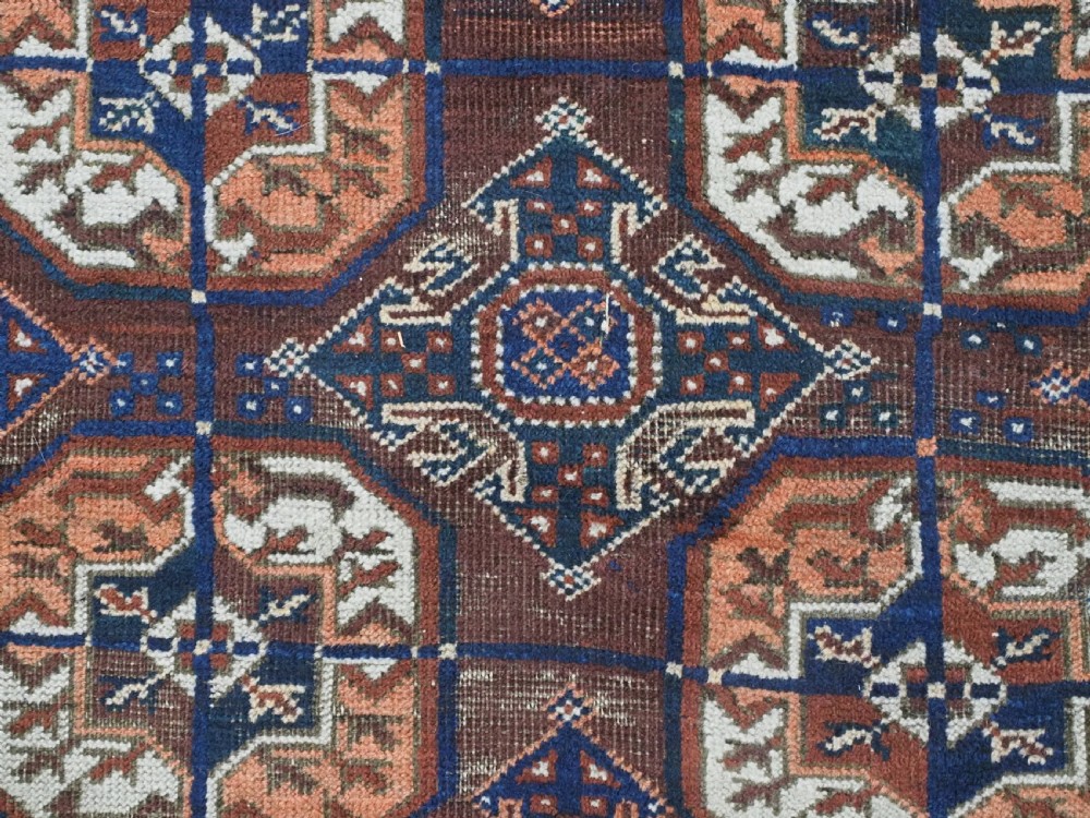 An antique hand-knotted Baluch wool rug featuring repeating geometric octagonal medallions in terracotta, navy, and ivory across a dark brown field with intricate multi-banded borders. View IX