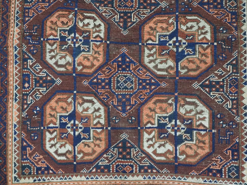 An antique hand-knotted Baluch wool rug featuring repeating geometric octagonal medallions in terracotta, navy, and ivory across a dark brown field with intricate multi-banded borders. View X