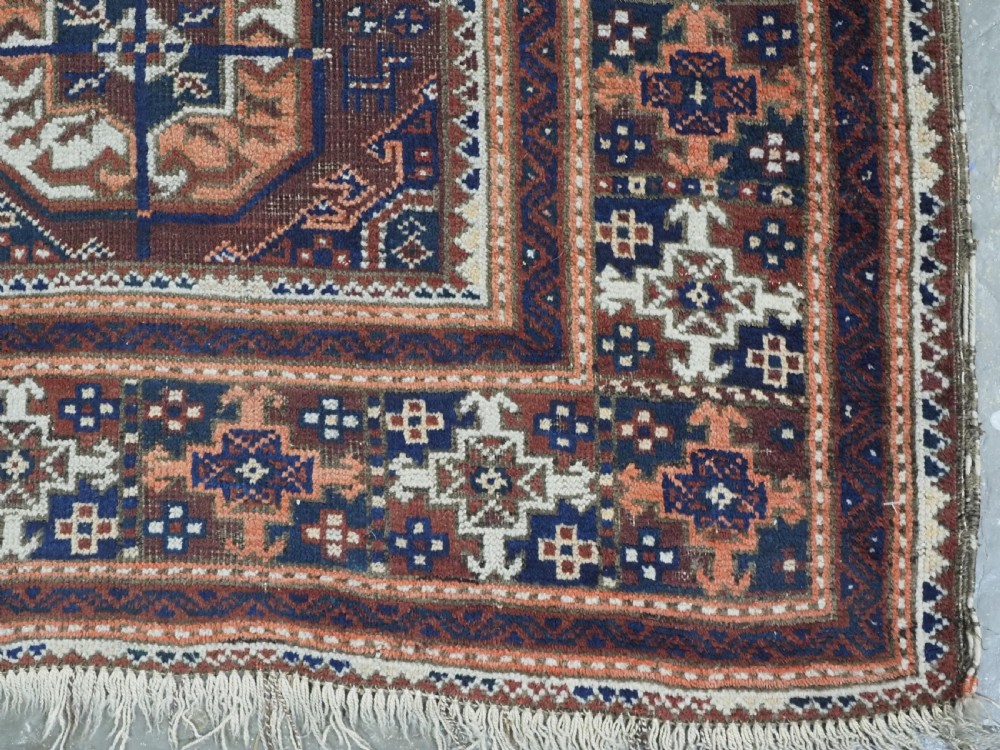 An antique hand-knotted Baluch wool rug featuring repeating geometric octagonal medallions in terracotta, navy, and ivory across a dark brown field with intricate multi-banded borders. View XI