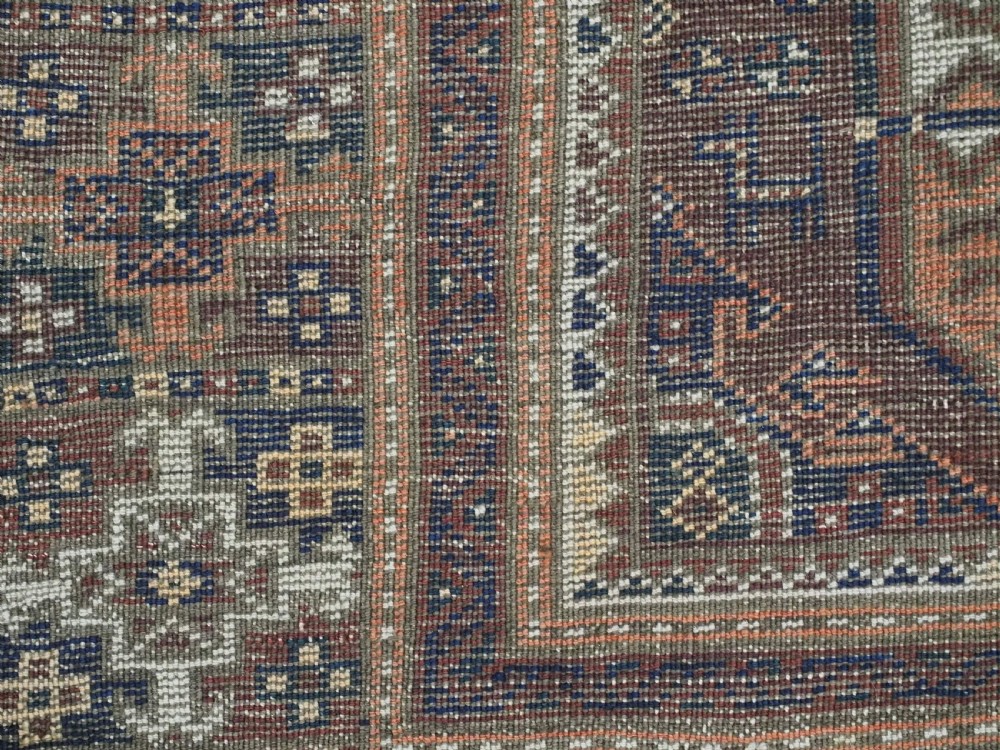 An antique hand-knotted Baluch wool rug featuring repeating geometric octagonal medallions in terracotta, navy, and ivory across a dark brown field with intricate multi-banded borders. View XII