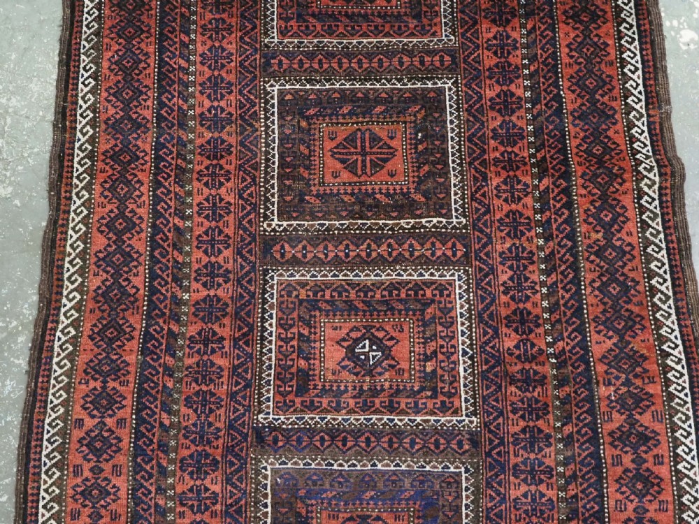 An antique Baluch hand-knotted wool rug featuring five geometric square medallions along a central field in deep red, dark navy blue, and terracotta tones. View IV