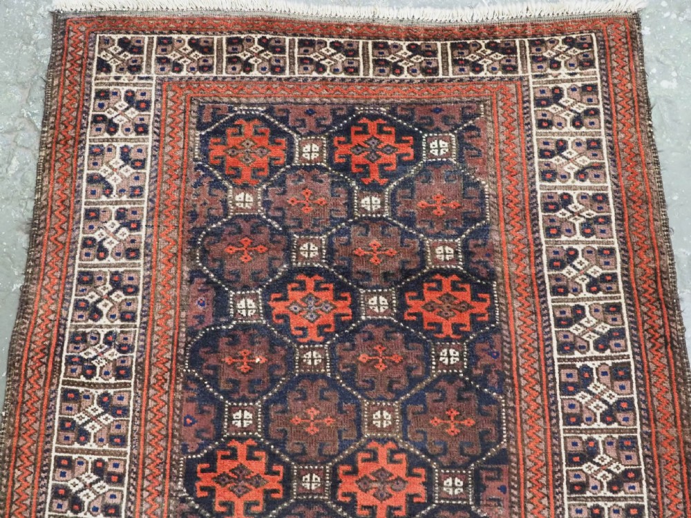 An antique rectangular Baluch wool rug featuring a dark central field with repeating geometric lattice patterns and a light ivory patterned border. View III