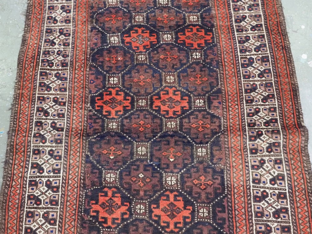 An antique rectangular Baluch wool rug featuring a dark central field with repeating geometric lattice patterns and a light ivory patterned border. View IV