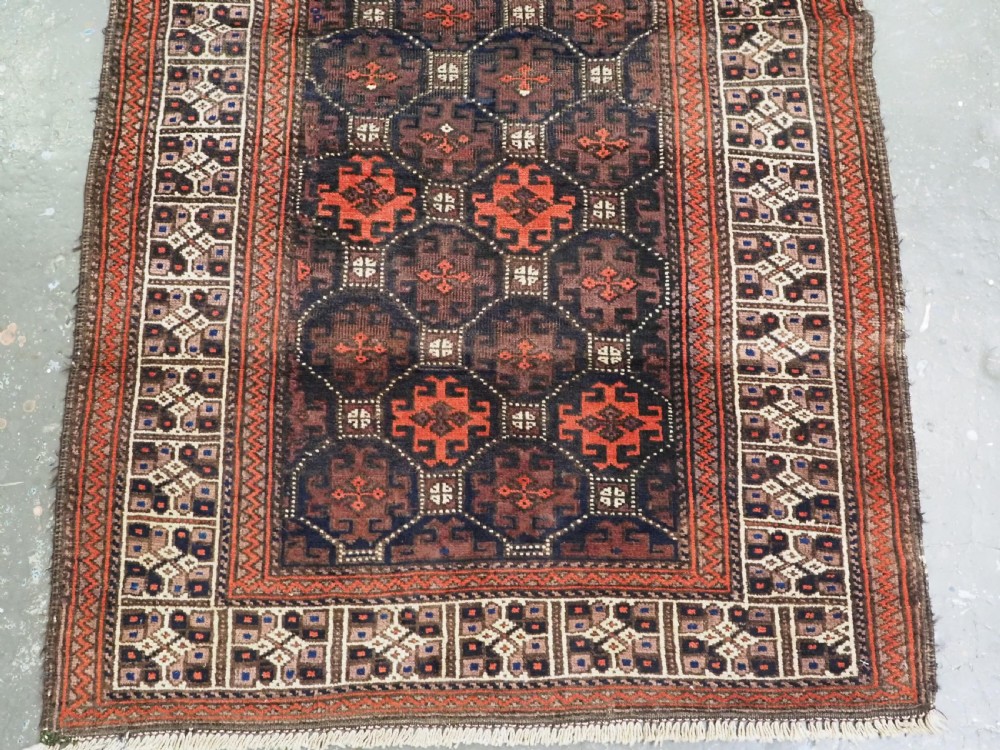 An antique rectangular Baluch wool rug featuring a dark central field with repeating geometric lattice patterns and a light ivory patterned border. View V