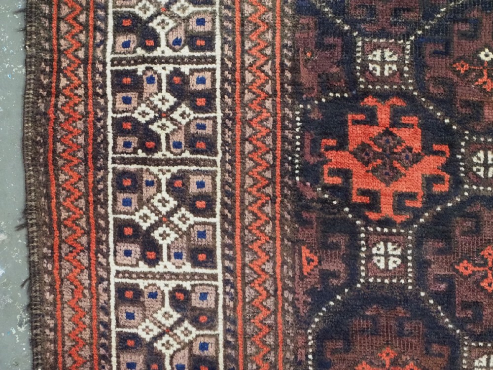 An antique rectangular Baluch wool rug featuring a dark central field with repeating geometric lattice patterns and a light ivory patterned border. View VII