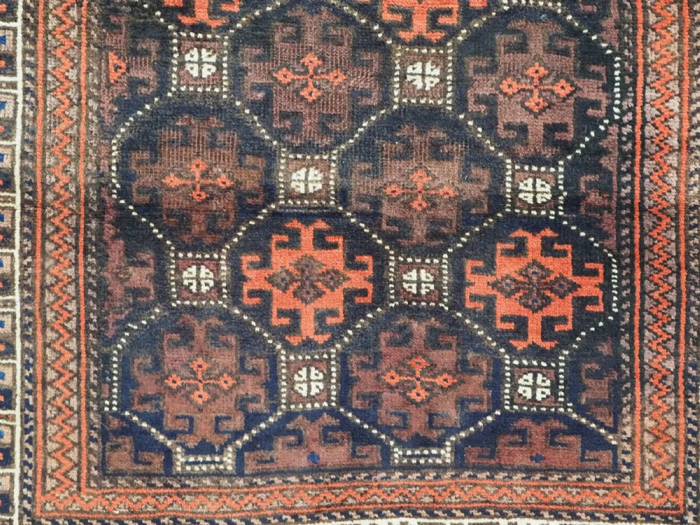 An antique rectangular Baluch wool rug featuring a dark central field with repeating geometric lattice patterns and a light ivory patterned border. View IX