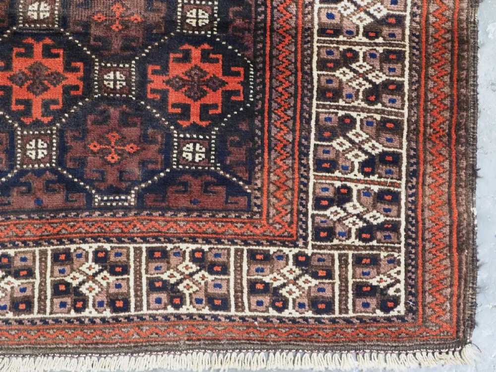 An antique rectangular Baluch wool rug featuring a dark central field with repeating geometric lattice patterns and a light ivory patterned border. View X