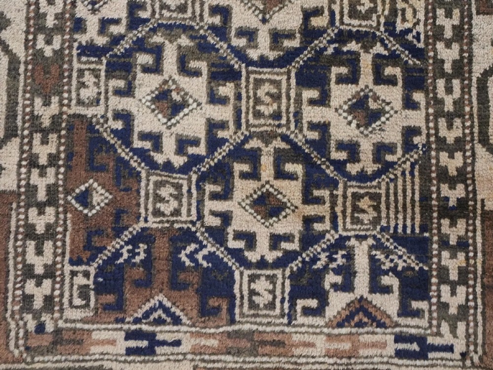 An antique woven Baluch rug featuring intricate geometric medallion patterns in shades of beige, brown, and dark indigo camel wool. View X