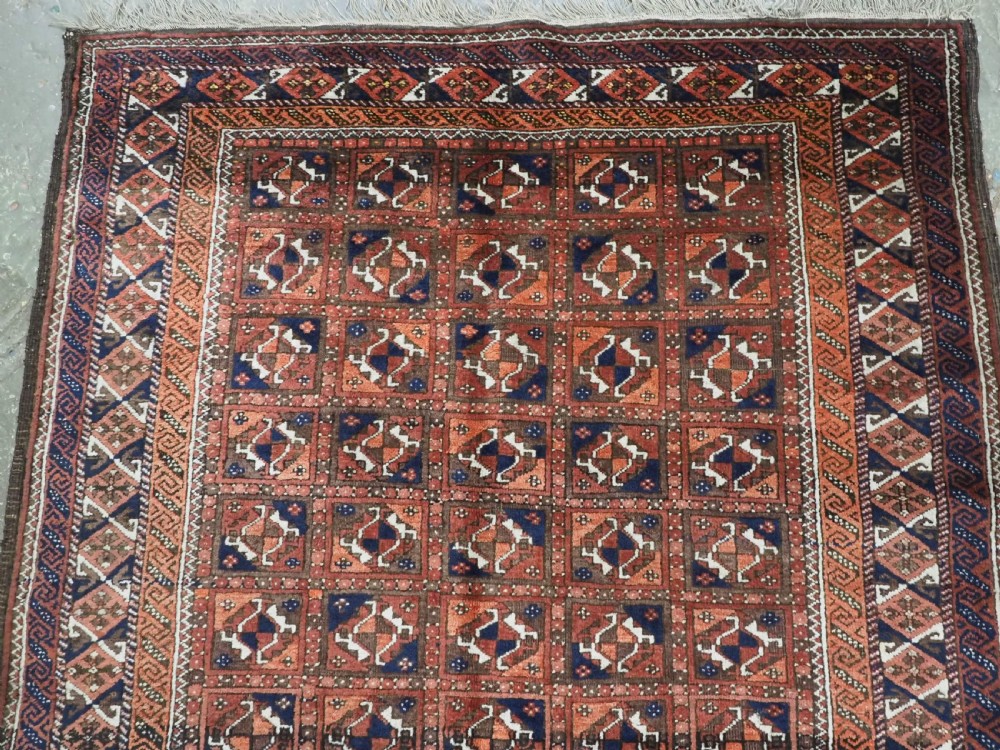 A rectangular hand-knotted antique Baluch rug featuring a grid of geometric motifs framed by decorative borders in terracotta, dark blue, and cream. View III