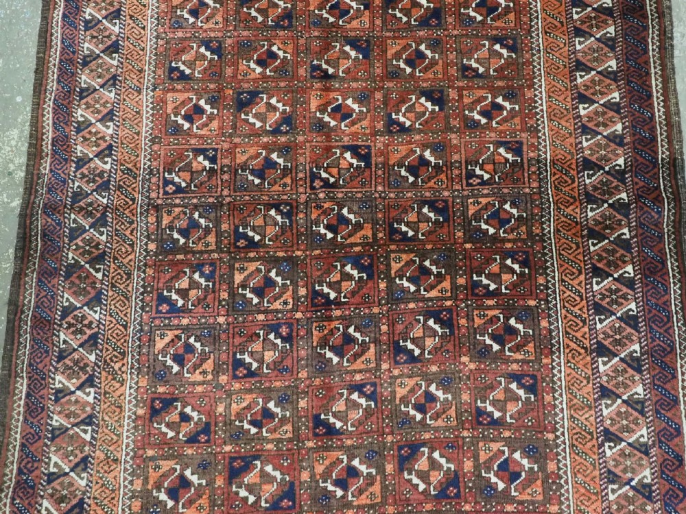 A rectangular hand-knotted antique Baluch rug featuring a grid of geometric motifs framed by decorative borders in terracotta, dark blue, and cream. View IV
