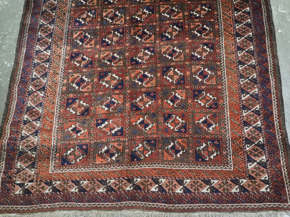 A rectangular hand-knotted antique Baluch rug featuring a grid of geometric motifs framed by decorative borders in terracotta, dark blue, and cream. View V