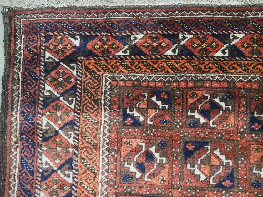 A rectangular hand-knotted antique Baluch rug featuring a grid of geometric motifs framed by decorative borders in terracotta, dark blue, and cream. View VI