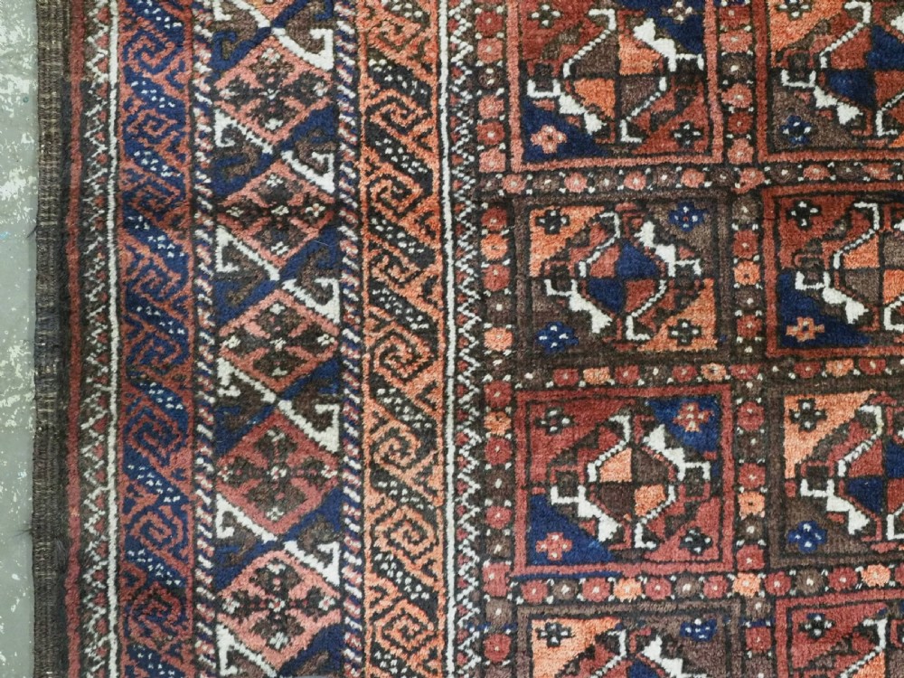 A rectangular hand-knotted antique Baluch rug featuring a grid of geometric motifs framed by decorative borders in terracotta, dark blue, and cream. View VII