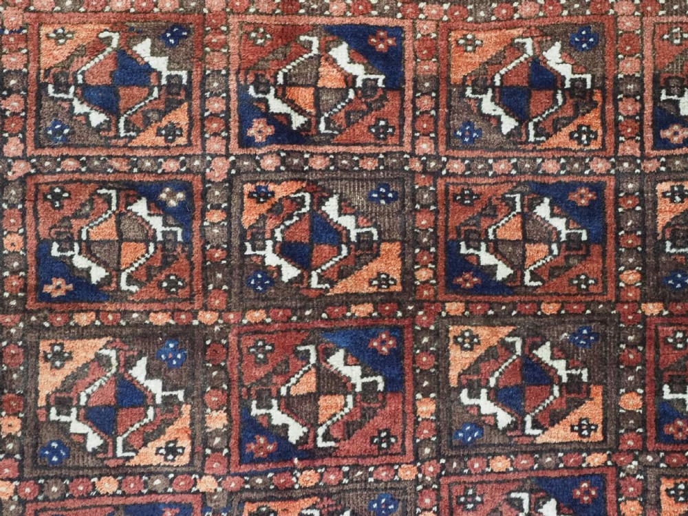A rectangular hand-knotted antique Baluch rug featuring a grid of geometric motifs framed by decorative borders in terracotta, dark blue, and cream. View IX