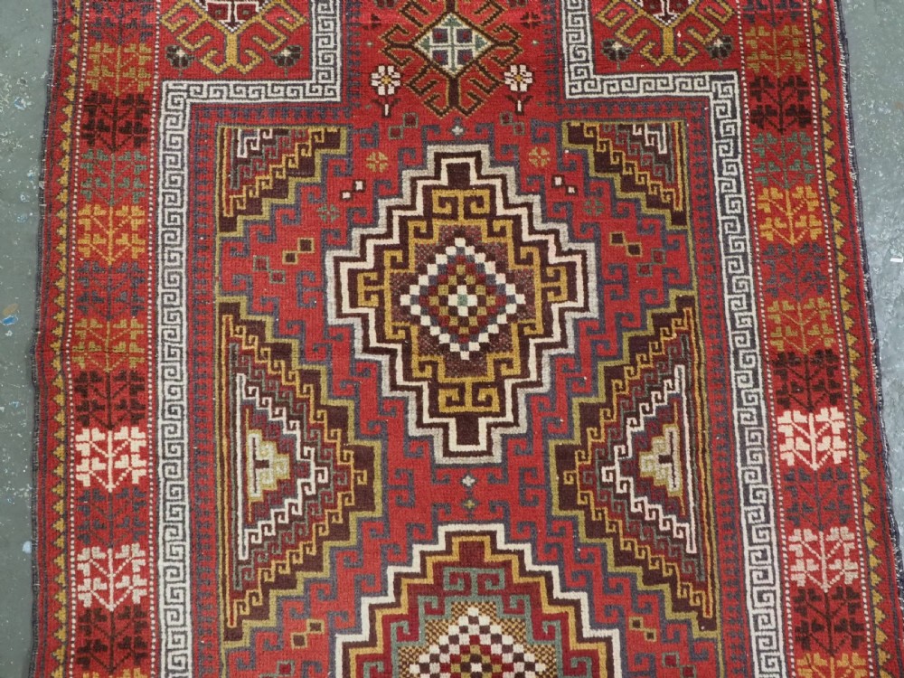 An antique hand-knotted Taimani Baluch prayer rug featuring a deep red field decorated with stepped geometric diamond motifs, a mihrab arch, and stylized geometric borders. View IV