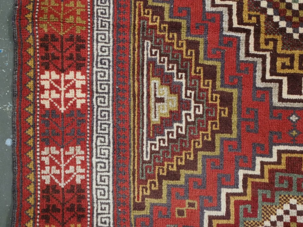 An antique hand-knotted Taimani Baluch prayer rug featuring a deep red field decorated with stepped geometric diamond motifs, a mihrab arch, and stylized geometric borders. View VI