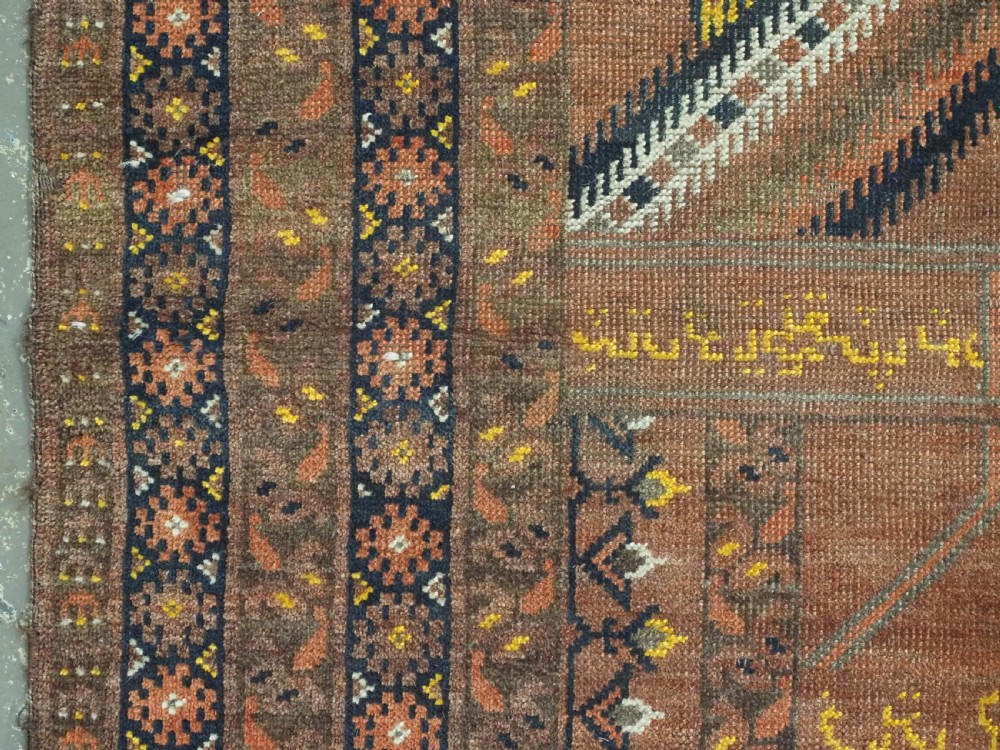 An antique hand-knotted Turkmen wool prayer rug featuring a terracotta-red field with geometric mihrab arches, woven script inscriptions, and patterned borders. View VII