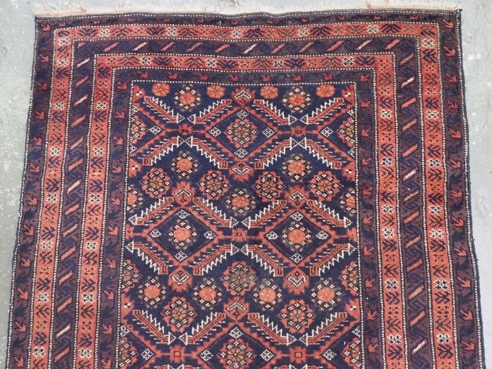 A hand-knotted antique Baluch wool rug featuring a dark navy central field filled with geometric diamond motifs framed by multi-tiered rust red borders. View III