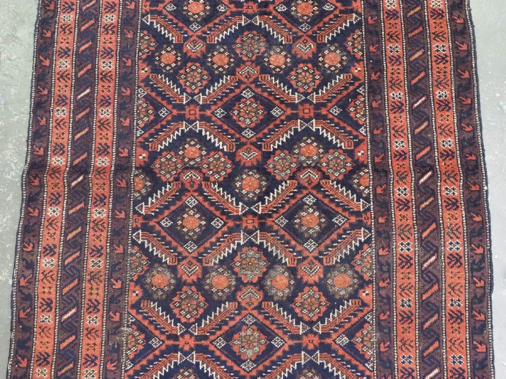 A hand-knotted antique Baluch wool rug featuring a dark navy central field filled with geometric diamond motifs framed by multi-tiered rust red borders. View IV