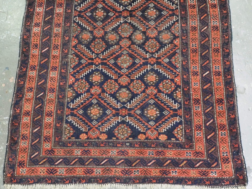 A hand-knotted antique Baluch wool rug featuring a dark navy central field filled with geometric diamond motifs framed by multi-tiered rust red borders. View V