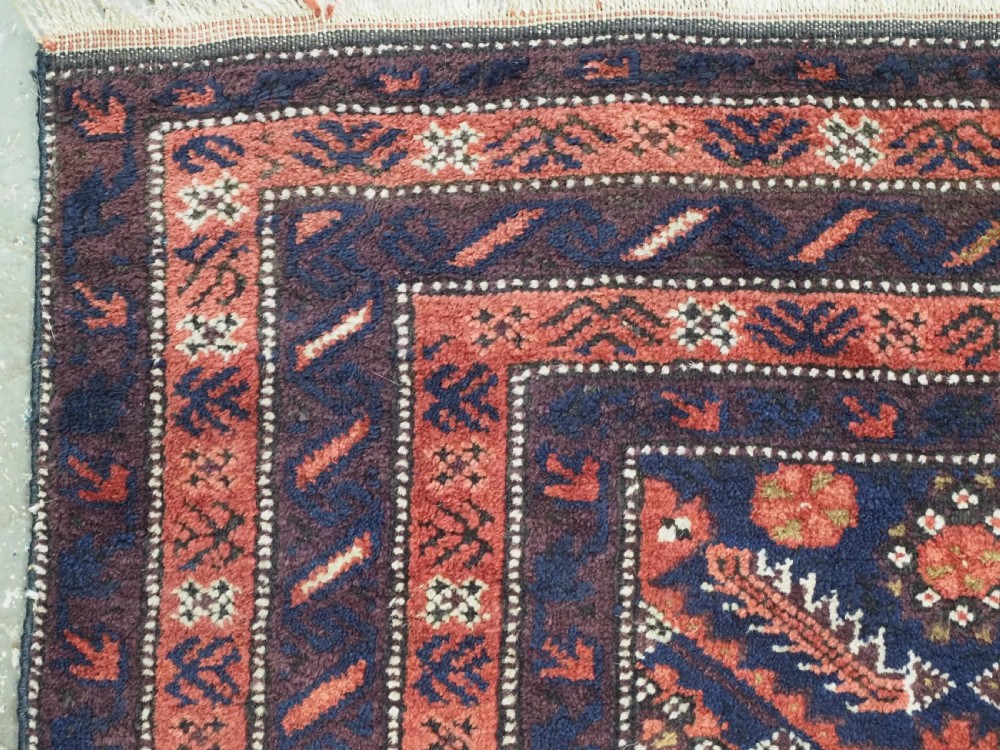 A hand-knotted antique Baluch wool rug featuring a dark navy central field filled with geometric diamond motifs framed by multi-tiered rust red borders. View VI
