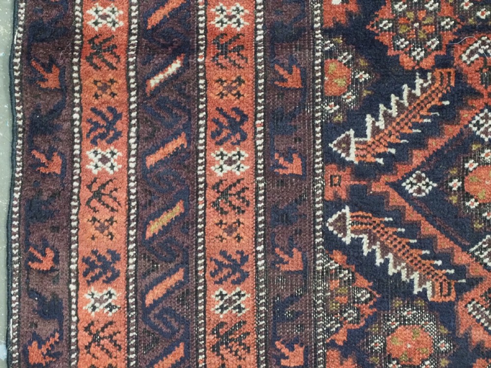 A hand-knotted antique Baluch wool rug featuring a dark navy central field filled with geometric diamond motifs framed by multi-tiered rust red borders. View VII