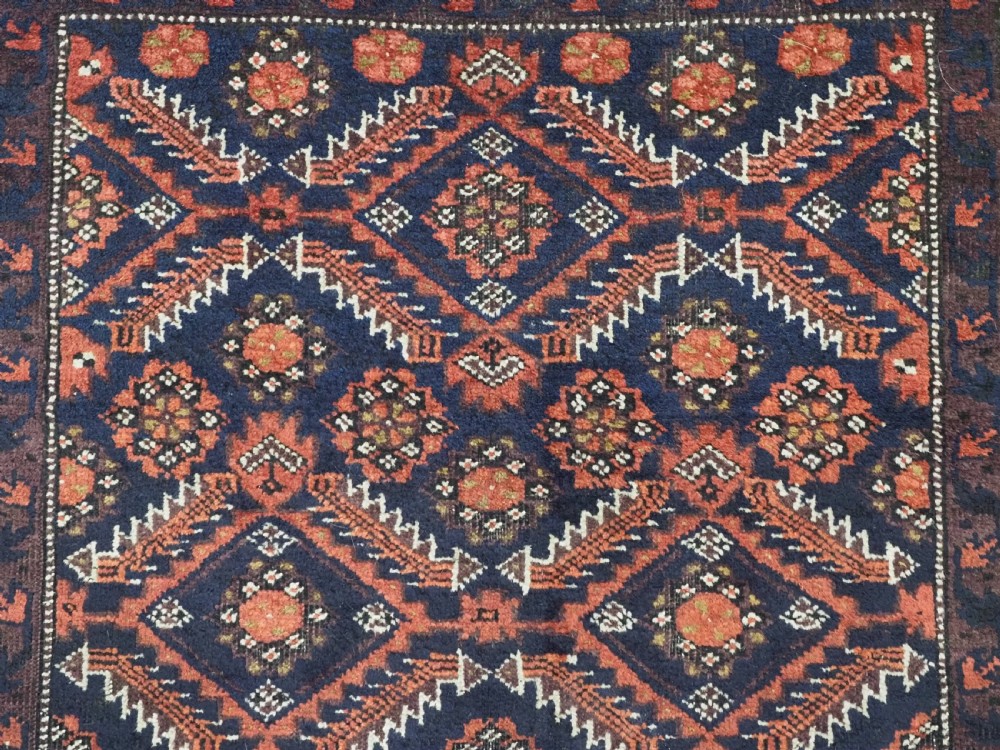 A hand-knotted antique Baluch wool rug featuring a dark navy central field filled with geometric diamond motifs framed by multi-tiered rust red borders. View VIII