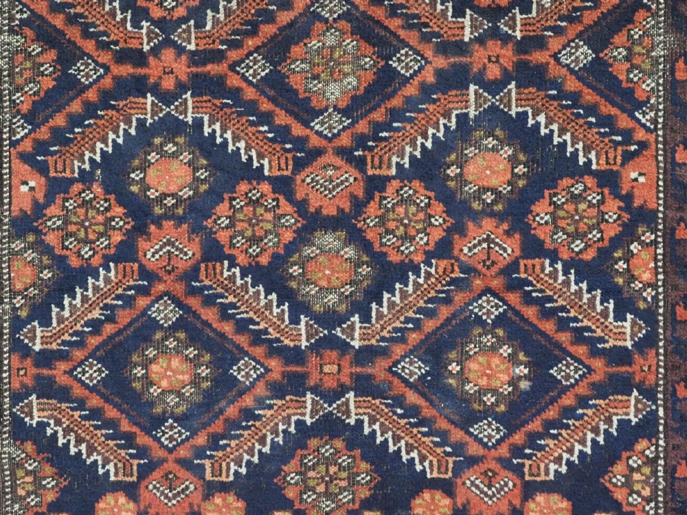 A hand-knotted antique Baluch wool rug featuring a dark navy central field filled with geometric diamond motifs framed by multi-tiered rust red borders. View IX