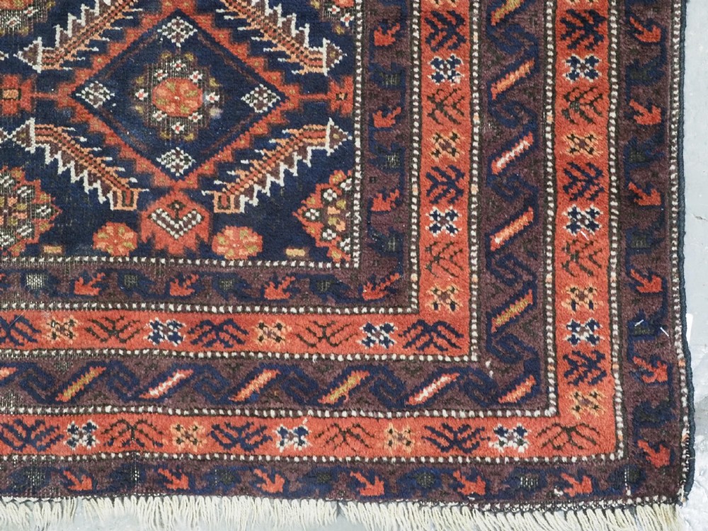 A hand-knotted antique Baluch wool rug featuring a dark navy central field filled with geometric diamond motifs framed by multi-tiered rust red borders. View X