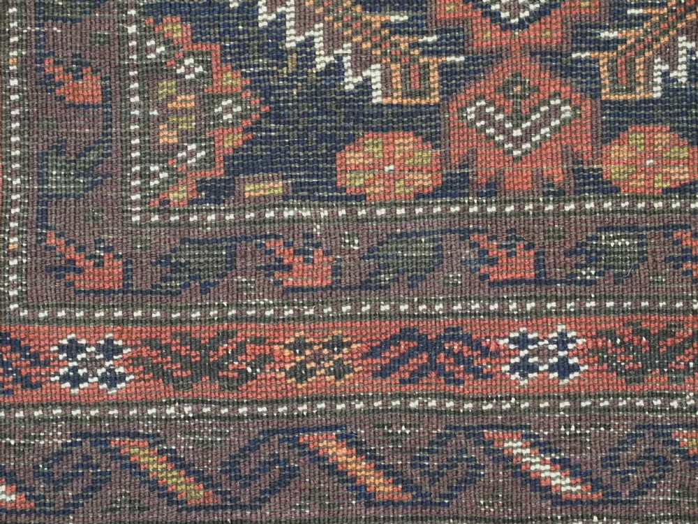 A hand-knotted antique Baluch wool rug featuring a dark navy central field filled with geometric diamond motifs framed by multi-tiered rust red borders. View XI