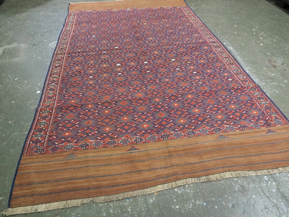 A rectangular handwoven wool kilim rug featuring a dense geometric diamond lattice pattern in terracotta red, navy blue, and cream with wide plain-woven border ends. View II