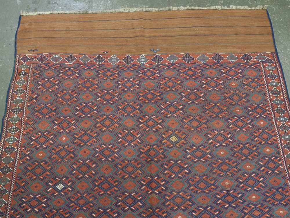A rectangular handwoven wool kilim rug featuring a dense geometric diamond lattice pattern in terracotta red, navy blue, and cream with wide plain-woven border ends. View III