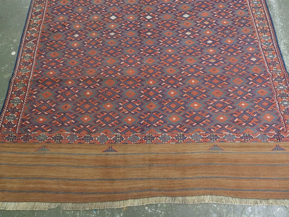 A rectangular handwoven wool kilim rug featuring a dense geometric diamond lattice pattern in terracotta red, navy blue, and cream with wide plain-woven border ends. View V