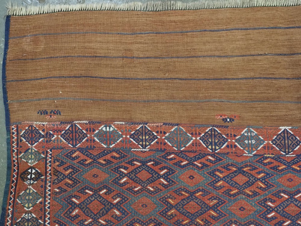 A rectangular handwoven wool kilim rug featuring a dense geometric diamond lattice pattern in terracotta red, navy blue, and cream with wide plain-woven border ends. View VI
