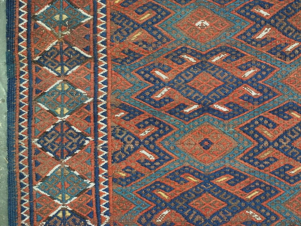A rectangular handwoven wool kilim rug featuring a dense geometric diamond lattice pattern in terracotta red, navy blue, and cream with wide plain-woven border ends. View VII